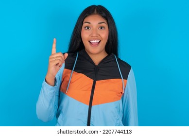 Pleasant Looking Young Latin Woman Wearing Sport Clothes Over Blue Background Has Clever Expression, Raises One Finger, Remembers Himself Not To Forget Tell Important Thing.