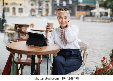 Pleasant mature lady relaxing at cafe terrace, using digital tablet and drinking coffee. Charming woman with grey hair enjoying free time on fresh air.