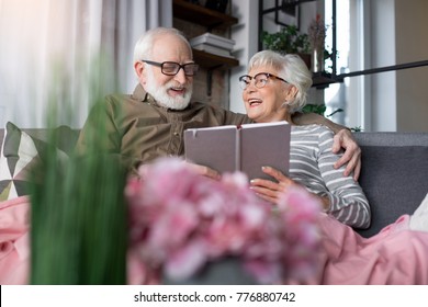 Pleasant Memories. Portrait Of Old Charming Couple Remembering Sweet Moments. Woman Is Looking At Husband While Laughing. Man Is Regarding At Picture While Tenderly Cuddling Wife