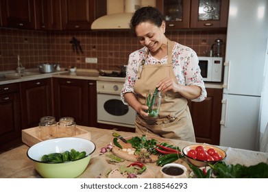 Pleasant middle-aged multiethnic woman wearing a kitchen apron, filling can with fresh chili peppers, making homemade pickles and marinated delicacies for the winter, in the home kitchen interior - Powered by Shutterstock