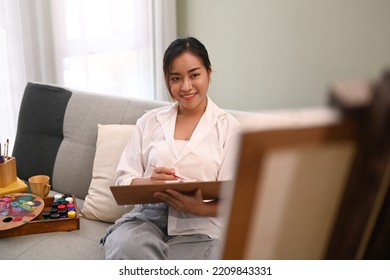 Pleasant Millennial Woman Sitting On Couch In Bright Living Room And Painting On Canvas. Art And Leisure Activity Concept