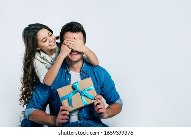 Pleasant Moment. Close-up Photo Of A Beautiful Little Girl With Long Hair Covering Eyes Of Her Dad In A Denim Jacket, While He Is Holding A Present With A Blue Ribbon On It.