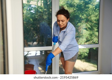 Pleasant Multi-ethnic Woman, Homemaker, Maid In Blue Work Rubber Gloves, A Lovely Housewife Wiping The Floor In A Veranda With Mop. Housekeeping. Domestic Chores. Housework. Cleaning Service.