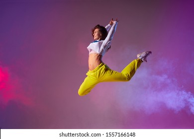 Pleasant playful woman with long curly brunette hair, bright yellow pants, white pullover, wearing stylish sneakers, showing dance element in club stage full of colourful smoke - Powered by Shutterstock