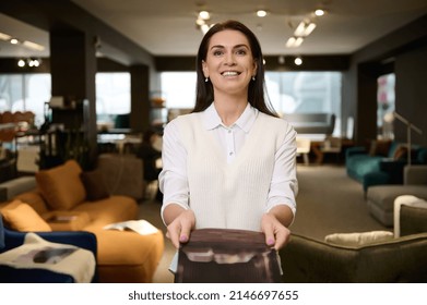 Pleasant Pretty Woman, Retail Assistant And Sales Rep Showing Catalog With Upholstered Furniture, Standing In The Exposition Center And Smiling With Cheerful Toothy Smile Looking At Camera