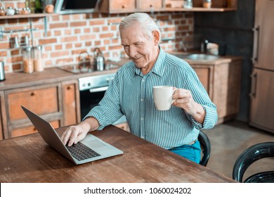Pleasant relaxation. Joyful delighted senior man sitting in front of the laptop and enjoying his drink while resting at home