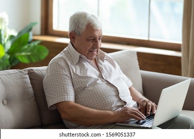 Pleasant Senior 70s Man Sitting On Sofa With Computer, Web Surfing Or Shopping Online. Happy Older Grandfather Communicating In Social Networks With Grown Up Children, Using Online Chat Application.