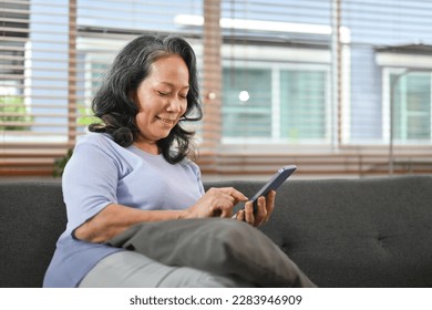 Pleasant senior woman sitting on couch browsing mobile apps, chatting with relatives online on mobile phone - Powered by Shutterstock