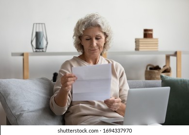 Pleasant Smiling Elderly Mature Woman Sitting On Sofa, Holding Laptop On Lap, Reading Banking Loan Paper Notification. Happy Older Senior Grandmother Looking Through Mail Letter Alone At Home.