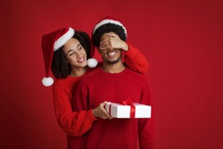 Pleasant Surprise For Soulmate Partner And Present At New Year And Xmas. Happy African American Lady In Santa Hat Closes Eyes Of Man In Sweater And Gives Gift, Isolated On Red Background, Studio Shot