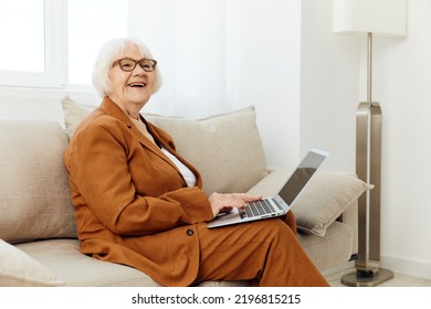 A Pleasant, Sweetly Smiling Elderly Old Lady Is Sitting With A Laptop On Her Lap On A Beige Sofa Dressed In A Stylish Brown Suit And Looking At The Camera