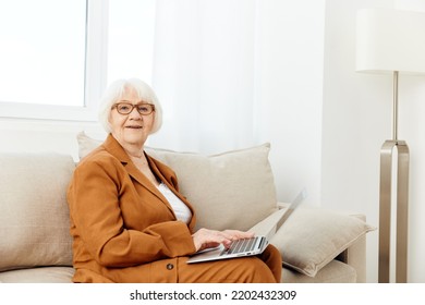 A Pleasant, Sweetly Smiling Elderly Old Lady Is Sitting With A Laptop On Her Lap On A Beige Sofa Dressed In A Stylish Brown Suit And Looking At The Camera