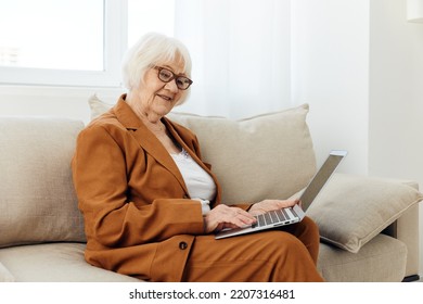 A Pleasant, Sweetly Smiling Elderly Old Lady Is Sitting With A Laptop On Her Lap On A Beige Sofa Dressed In A Stylish Brown Suit And Looking At The Camera