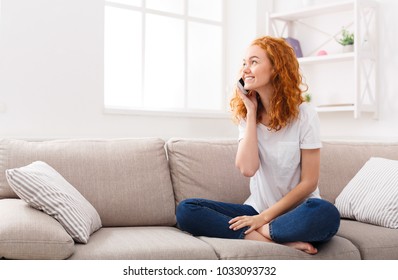 Pleasant talk with friend. Attractive smiling young redhead woman talking on cell phone while sitting on the beige sofa at home. Technology, communication and coziness concept