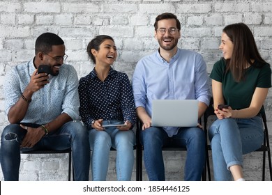 Pleasant waiting. Group of four multiethnic millennial men and women teammates friends spending time in queue at wifi zone talking laughing sharing information by internet app using electronic devices