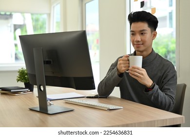 Pleasant young asian man freelancer drinking hot coffee and looking at computer screen - Shutterstock ID 2254636831