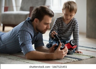 Pleasant Young Father Lying On Floor Carpet, Imagining Repairing Car With Focused Little Kid Son. Small Child Boy Holding Screwdriver, Fixing Broken Toy With Controlling Dad, Skills Transmission.