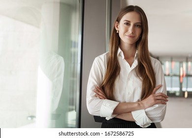 Pleasant Young Female Assistant, Manager Standing In Office Corridor Near Window, With Pleased Confident Expression, Cross Hands Over Chest Showing Readiness, Business And Women Concept