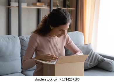 Pleasant young female client unpacking huge cardboard box, sitting on cozy couch at home, satisfied with fast delivery service. Happy millennial woman unboxing parcel with order from internet store.