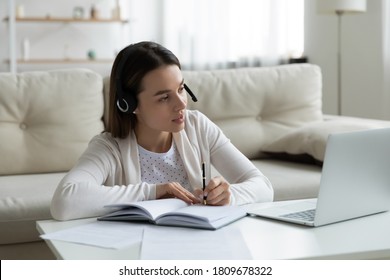 Pleasant young female PhD student in headphones sitting at coffee table in living room watching educational lecture, attending online courses webinar, consulting with supervisor about thesis writing.