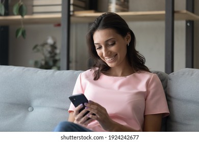 Pleasant young indian female relax at home with cellphone look at screen read awaited email message buy order goods services online. Cheerful multiracial woman sit on sofa play mobile game at phone app