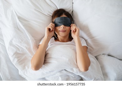Pleasant young woman laying in bed and putting eye mask on before going to sleep