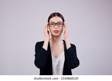 Pleasant-looking Business Woman Looking Straight And Is Standing On A Gray Background In A Black Jacket, White T-shirt And Computer Glasses. The Girl's Hands Holds The Edges Of The Glasses. Worker.