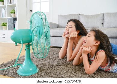 Pleasantly Mother With Young Little Daughter Lying Down On Living Room Floor And Face To Electric Fan Enjoying Blowing Cool Wind Together At Summer.