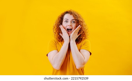 Pleasantly Surprised Young Woman With Curly Hair Ginger Standing With Gaping Mouth, Hands On Her Face. Over Yellow Background. Wearing An Orange Shirt.