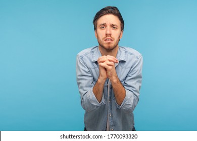 Please, I'm begging! Portrait of unhappy man in worker denim shirt keeping hands in prayer gesture and looking with imploring expression, sincere asking permission. indoor studio shot, blue background