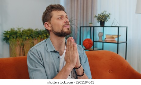 Please, God Help. Man Praying Sincerely With Folded Arms, Looking Upward And Making Wish, Asking With Hopeful Imploring Expression, Begging Apology. Young Guy Sitting On Couch At Home Living Room