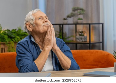 Please, God help. Worried senior man praying for heath sincerely with folded arms, looking upward making wish, asking hopeful imploring expression begging apology at home. Elderly mature grandfather - Powered by Shutterstock