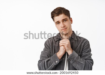 Please help me. Cute smiling young man, coy guy begging you, holding hands in pray and looking silly as asking for favour, need advice, white background