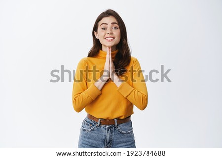 Please help me. Cute young woman asking for favour, need advice or favour, holding hands in pray gesture and smiling, standing against white background.