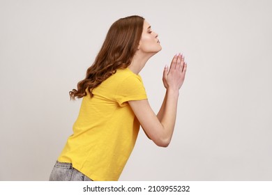 Please help me or forgive. Side view of worry teenager girl with wavy hair in casual yellow T-shirt standing with palm hands, pleading. Indoor studio shot isolated on gray background. - Shutterstock ID 2103955232