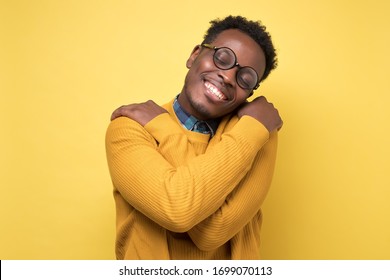 Pleased african american man in yellow clothes and glasses hugs himself, has high self esteem. Studio shot on colored wall. - Shutterstock ID 1699070113