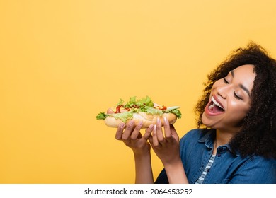 pleased african american woman eating tasty hot dog isolated on yellow