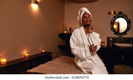 Pleased african girl sitting on massage table and touching herself in spa salon. Young attractive woman in bathrobe and wrapped bath towel on head with closed eyes. Concept of rest with spa treatment - Powered by Shutterstock