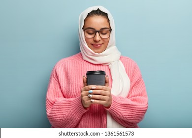 Pleased Arabic lovely woman warms hands on hot paper cup with drink, enjoys morning coffee, has gentle friendly expression, wears white scarf on head, loose pink jumper, poses indoor. Spare time