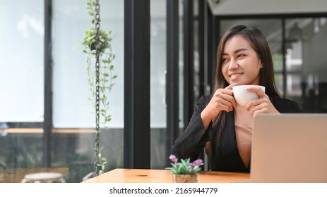 Pleased asian businesswoman enjoying morning coffee and looking through a window at office - Shutterstock ID 2165944779