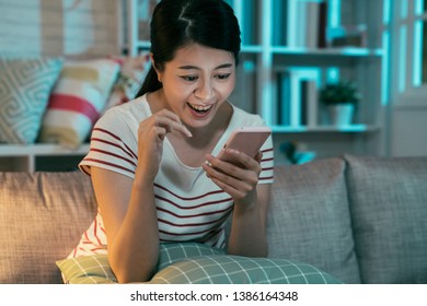 Pleased Asian Chinese Woman In T-shirt Writing Message On Smartphone In Modern Interior House At Night. Young Girl Stay Up Late Sitting On Couch Using Mobile Phone Laughing Chatting With Friend.