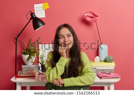 Pleased Asian girl sits against desktop with all necessary supplies for studying, smiles and daydreams during break, poses on chair against bright rosy wall, sticky notes with inscription. Education