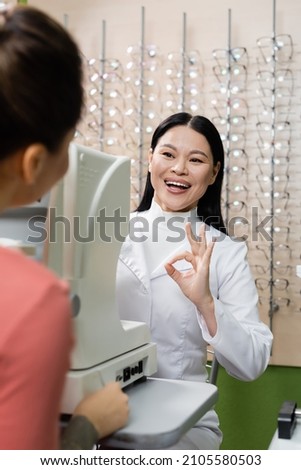 pleased asian oculist showing okay gesture near vision screener and blurred woman in optics salon