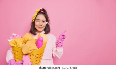 Pleased Asian Woman Housekeeper With Dark Hair Wears Sweatshirt And Protective Rubber Gloves Holds Laundry Basket Indicates Aside On Blank Space Isolated Over Pink Background. Housecleaning Concept