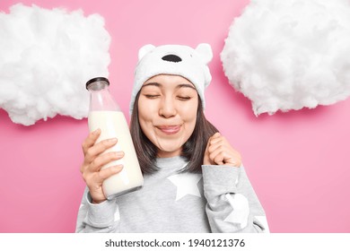 Pleased Asian Woman Licks Lips Closes Eyes Wants To Drink Milk For Breakfast Dressed In Domestic Comfortable Clothes Raises Hand Feels Temptation Isolated Over Pink Background White Clouds Above