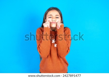 Pleased beautiful kid girl wearing orange knitted sweater with closed eyes keeps hands near cheeks and smiles tenderly imagines something very pleasant