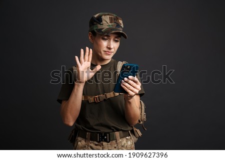 Pleased beautiful soldier woman waving hand while using cellphone isolated over black background