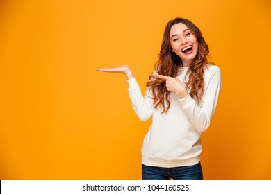 Pleased brunette woman in sweater holding copyspace on the palm and pointing on it while looking at the camera over yellow background