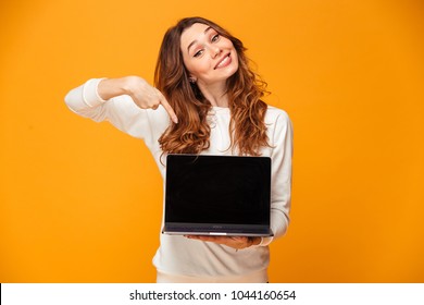 Pleased Brunette Woman In Sweater Showing Blank Laptop Computer Screen And Pointing On It While Looking At The Camera Over Yellow Background