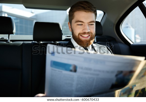 Pleased business man reading newspaper while sitting\
on back seat of a car